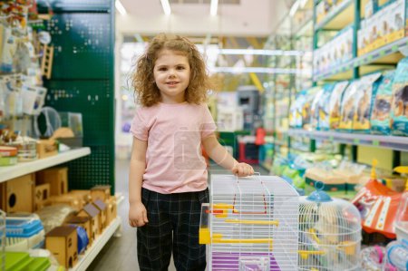 Téléchargez les photos : Portrait de petite fille enfant debout à proximité cage pour rongeur à animalerie. Choisir la maison pour l'animal domestique - en image libre de droit