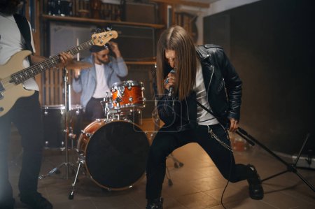 Photo for Expressive rocker singing song in microphone while his band playing heavy metal on drum and electric bass guitar. Young guys at rehearsal repetition in home studio - Royalty Free Image