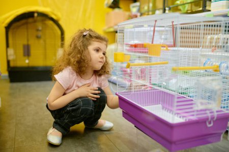 Foto de Niña pequeña sentada cerca de la jaula para roedores en la tienda de mascotas en busca de un nuevo hogar para su adorable animal doméstico - Imagen libre de derechos