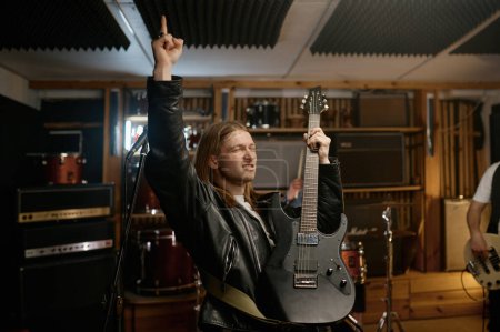 Photo for Portrait of expressive rocker playing last chord of song on his guitar raising hand with index finger up. End of performance of rock band - Royalty Free Image