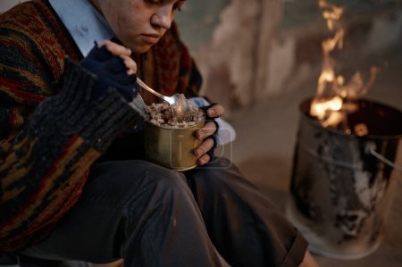 Foto de Pobre mujer hambrienta sin hogar comiendo conservas de alimentos enlatados mientras se calienta en llamas de hoguera. Disparo de cosecha - Imagen libre de derechos