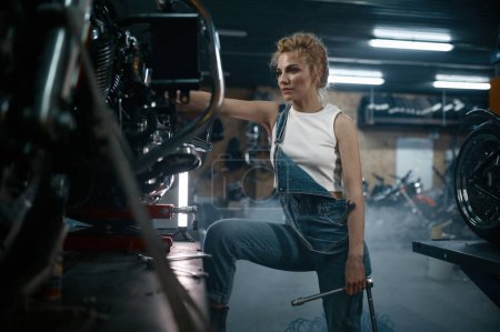 Photo for Talented girl wearing blue jumpsuit using work tool to repair motorcycle in garage. Pensive young woman at creative authentic workshop - Royalty Free Image