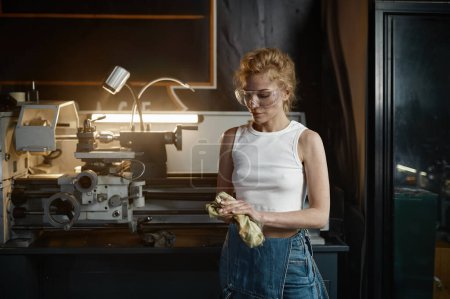 Photo for Young woman wearing denim overalls and protective safety goggles wiping her hands with rag after working on lathe turning machine at workshop - Royalty Free Image