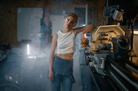 Photo for Young woman wearing denim overalls and protective safety goggles after working on lathe turning machine at workshop - Royalty Free Image