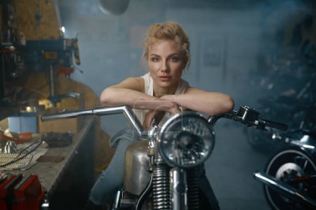 Téléchargez les photos : Portrait de belle femme motard assis sur la moto dans le garage. Jolie femme mécanicienne posant pour caméra à l'atelier - en image libre de droit