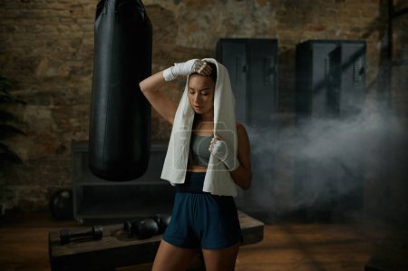 Téléchargez les photos : Fatigué sportif attrayant boxer femme portant une serviette sur la tête et des bandages de kickboxing debout à la salle de gym prendre une pause après l'entraînement dur - en image libre de droit