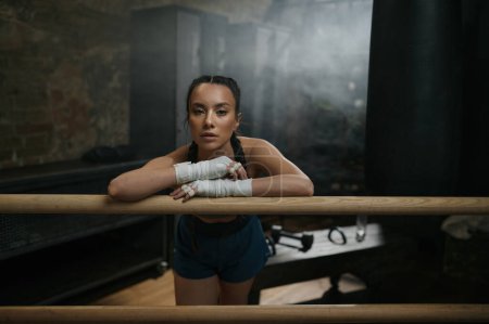 Photo for Pretty young woman boxer resting at boxing ring ready for workout. Tired female fitness athlete exercising and relaxing before or after strong sports training - Royalty Free Image