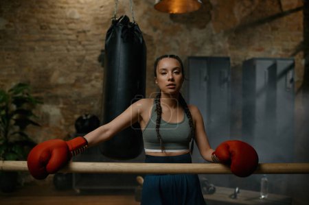 Photo for Athletic female fighter with red boxing gloves standing leaned at wooden training bar in gym with loft interior design - Royalty Free Image