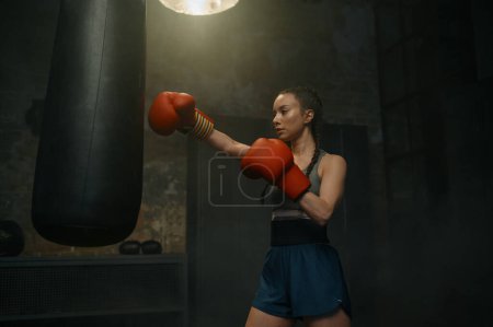 Foto de Fuerte boxeo femenino con saco de boxeo que tiene entrenamiento de entrenamiento. Mujer ejercitando la práctica de defensa personal en el gimnasio - Imagen libre de derechos