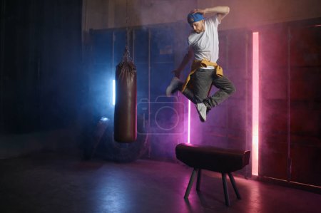 Foto de Joven adulto b-boy breakdancer saltando en el aire sobre el fondo del gimnasio loft en luz de neón. Guapo hipster hombre haciendo acrobacia realizar breakdance freestyle - Imagen libre de derechos
