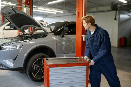 Foto de Mecánico automático masculino empujando caja de herramientas con ruedas para el trabajo en la estación de servicio de reparación de automóviles. Ocupación y empleo - Imagen libre de derechos