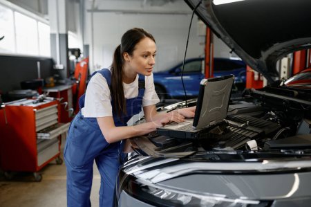 Photo for Young woman car engineer providing computer diagnostics using laptop with special interface - Royalty Free Image