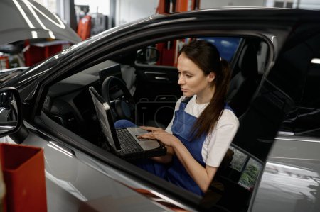Photo for Young woman technician sitting in car cabin working on laptop providing computer diagnostics. Auto service station - Royalty Free Image