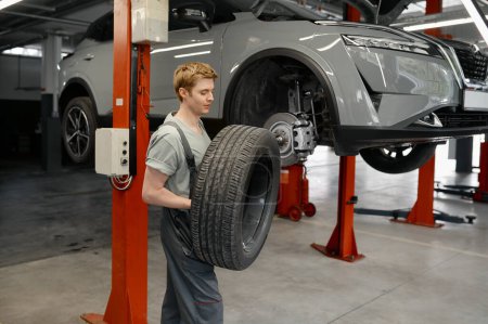 Foto de Serious joven mecánico de automóviles que lleva el neumático para el reemplazo. Servicio de reparación profesional y reparación de automóviles - Imagen libre de derechos