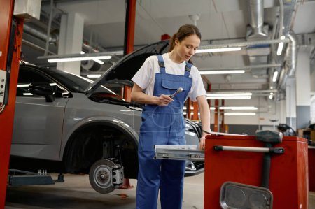 Photo for Young woman auto mechanic taking instrument for work from toolkit box for car repair in mechanical workshop - Royalty Free Image