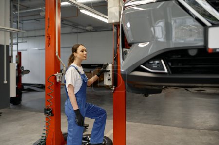 Photo for Young woman auto technician turning on car lift mechanism while working at repair workshop - Royalty Free Image