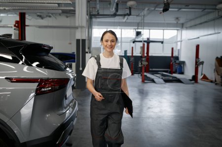 Téléchargez les photos : Jeune femme mécanicien avec presse-papiers près de la voiture à l'atelier de service automobile - en image libre de droit