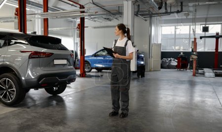 Téléchargez les photos : Jeune femme mécanicien écrit à la liste de contrôle presse-papiers pour la réparation de voiture de travail dans l'atelier de garage - en image libre de droit