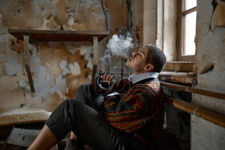 Foto de Mujer sin hogar fumando sentado en el piso de la habitación de la casa abandonada y mirando a la cámara - Imagen libre de derechos