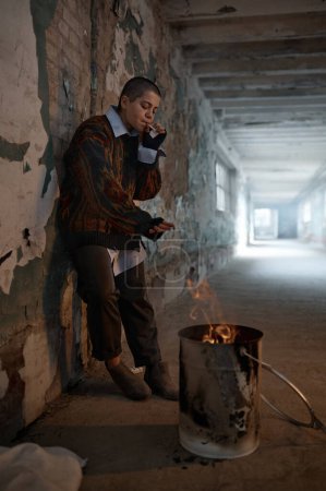 Foto de Pobre adolescente sin hogar fumando mientras se calienta en hoguera en barril sobre la escena de la casa de refugio abandonado urbano - Imagen libre de derechos