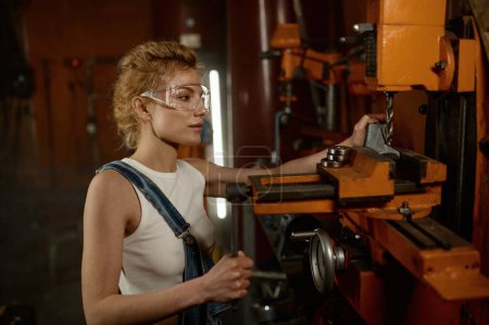 Téléchargez les photos : Vue rapprochée d'une jeune femme professionnelle sérieuse et concentrée portant des lunettes de protection de sécurité travaillant sur une fraiseuse électrique et une perceuse dans un atelier de garage - en image libre de droit
