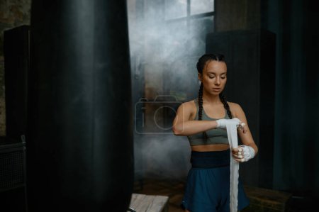 Téléchargez les photos : Gros plan atmosphérique de boxeuse portant un survêtement enveloppant ses mains dans un bandage debout à proximité d'un sac de boxe dans un ancien club de sport vintage - en image libre de droit