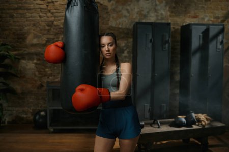 Foto de Joven boxeadora confiada que usa guantes descansa cerca del saco de boxeo en el viejo gimnasio deportivo loft - Imagen libre de derechos