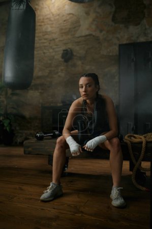 Téléchargez les photos : Jeune boxeuse sportive en bandage de boxe assise sur un banc au-dessus de l'intérieur de la salle de gym loft. Kickboxing club d'entraînement pour femme - en image libre de droit