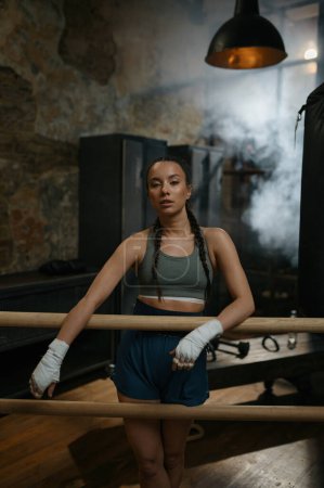 Photo for Athletic female fighter with bandaged hands standing leaned at wooden training bar in the loft gym interior - Royalty Free Image