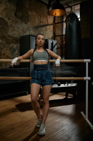 Photo for Athletic female fighter with bandaged hands standing leaned at wooden training bar in gym with loft interior design - Royalty Free Image