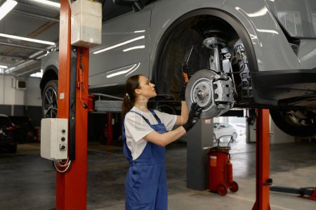 Foto de Mujer mecánico automático cubo de la rueda de fijación o freno de disco de coche levantado en el mecanismo de elevación utilizando llave inglesa. Trabajo en el servicio de garaje - Imagen libre de derechos