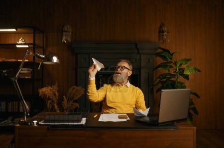 Photo for Serious thoughtful senior man throwing paper plane while sitting at work table with laptop computer and origami ship in home office. business startup launch, break time, dreaming about travelling - Royalty Free Image