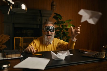 Foto de Divertido hombre maduro lleno de alegría con gafas piloto jugando con el avión de origami mientras está sentado en el lugar de trabajo en la oficina en casa por la noche - Imagen libre de derechos
