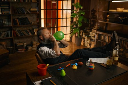 Foto de Relajado hombre de negocios senior con ropa elegante inflando globo mientras está sentado en la oficina en casa - Imagen libre de derechos