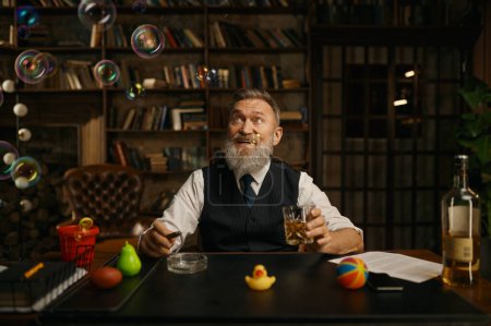 Foto de Retrato de un hombre de negocios mayor relajado fumando cigarro y bebiendo whisky mientras está sentado en la mesa de trabajo en la oficina en el hogar. Mirando burbujas volando en el aire - Imagen libre de derechos