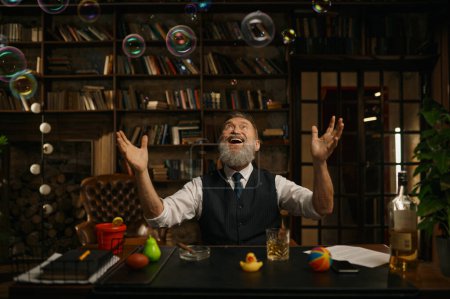 Foto de Feliz alegre y juguetón empresario sénior divertido sentado en la mesa de escritorio bajo el ventilador de burbujas regocijándose y sintiendo alegría - Imagen libre de derechos