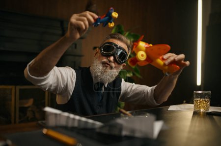 Foto de Juguetón jefe jugando avión de juguete en la mesa de trabajo. Maduro hombre de negocios de pelo gris con ropa elegante y gafas piloto de avión divertirse y relajarse - Imagen libre de derechos