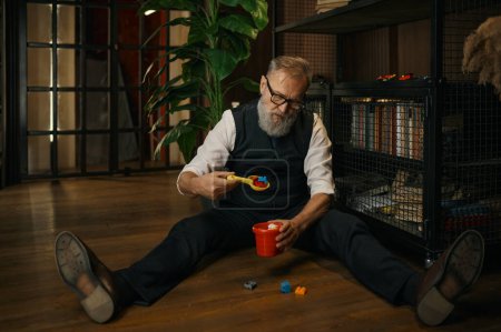 Téléchargez les photos : Homme d'affaires senior drôle jouant panier jouet, scoop et blocs de construction tout en étant assis sur le sol dans le bureau à domicile de luxe - en image libre de droit