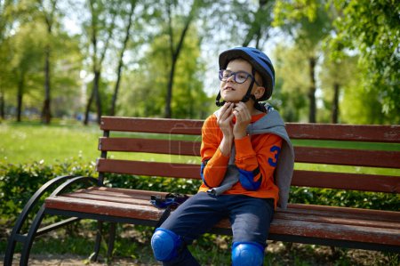 Photo for Little boy wearing protective helmet on head while sitting on bench in park. Preparation for safety extreme riding roller skates outdoors and active lifestyle concept - Royalty Free Image