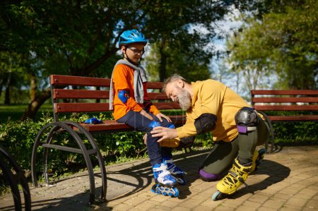 Photo for Grandfather helping grandson to put on roller skates and protective gear. Happy family active rest and leisure recreation time in urban park on weekend - Royalty Free Image