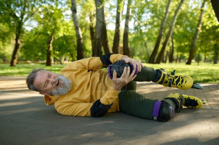 Foto de Hombre mayor sintiendo dolor en la rodilla por una lesión al caer mientras patina en el parque. Accidente y accidente mientras patinaba - Imagen libre de derechos
