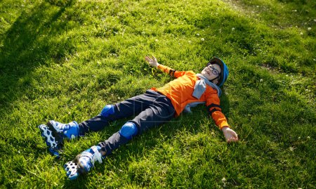 Photo for Happy tired little preschool boy wearing roller skates and helmet rest on green grass in urban park - Royalty Free Image