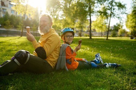 Téléchargez les photos : Fatigué et heureux grand-père et petit-fils ayant de la glace après le patinage à roulettes tout en étant assis dos à dos sur le champ de rue herbe verte - en image libre de droit