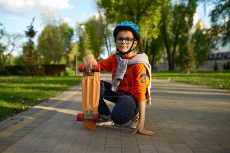 Foto de Retrato de un colegial sonriente con casco protector sentado en asfalto con monopatín. Niño pequeño atleta tiro de cuerpo entero. Deporte de seguridad para niños - Imagen libre de derechos