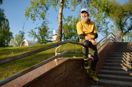 Photo for Happy smiling senior man wearing roller skates rest sitting on stair railing in urban park. Active time on retirement - Royalty Free Image