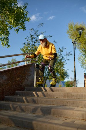 Foto de Valiente hombre mayor montando patines moviéndose por las escaleras del parque haciendo truco peligroso. Deporte extremo para personas mayores - Imagen libre de derechos