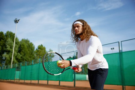 Téléchargez les photos : Joueur de tennis masculin concentré avec raquette et entraînement au ballon sur un terrain extérieur. Préparation pour le service - en image libre de droit