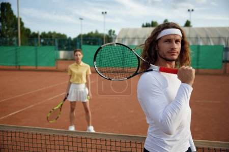Téléchargez les photos : Des amis jouant au tennis sur le terrain. Concentrez-vous sur l'homme confiant joueur tenant raquette sur l'épaule et regardant à la caméra. Week-end loisirs sportifs pour les jeunes couple - en image libre de droit
