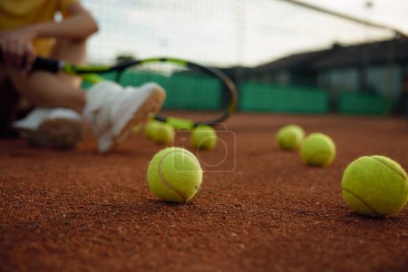 Téléchargez les photos : Repos après le match tourné avec l'accent sur les balles dispersées sur le terrain de jeu et joueur de tennis détendu assis sur le sol - en image libre de droit