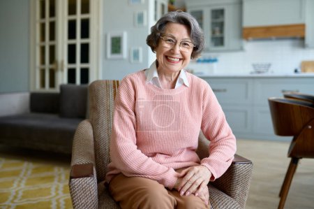 Foto de Retrato de una feliz anciana sonriente mirando a la cámara mientras está sentada en una silla en su propia sala de estar - Imagen libre de derechos
